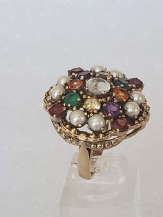 "I'm excited to have come across this beautiful ring because it is in a bigger size and has lots of jewels. The metal is gold plated with a large undercarriage with holes. Each stone is prong set and are in different colored gemstones. The ring has a hallmark \"NB NICKY BUTLER\" and has its original ring box. All stones are intact with non-missing. The ring was used a couple of times and then put away. The finish is shiny with no scratches or other oddities. Makes a great cocktail ring for the H Antique Gold Cluster Ring With Multi-stone, Antique Multi-stone Cluster Jewelry, Antique Multicolor Multi-stone Jewelry, Vintage Gold Multi-stone Cluster Ring, Vintage Multi-stone Pearl Ring For Anniversary, Heirloom Multi-stone Pearl Ring Gift, Heirloom Multi-stone Pearl Ring As Gift, Unique Jeweled Rings For Gift, Heirloom Multicolor Ring Jewelry