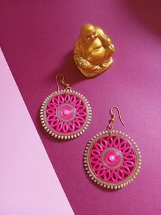 two pairs of pink and green earrings sitting on top of a purple table next to a gold buddha statue