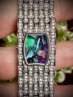 Gemstone-filled shaker locket up-cycled designer wrist watch, silver tone with pave rhinestone, opal, amethyst, aquamarine, topaz, unique gift for her. A kaleidoscope on your wrist! Formerly a blingy wrist watch, inlaid with pave rhinestones on the top of the wrist. Now, minus the mechanism, I've filled the space with a collection of colourful gems. Most notable is a large chunk of raw Ethiopian opal in a gorgeous deep iridescent green. A constantly shifting mix of blue, purple, green, and a few Iridescent Green, Unique Gifts For Her, Purple Green, Chain Link Bracelet, Ethiopian Opal, Link Bracelets, Blue Purple, Chain Link, Aquamarine