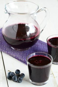 two glasses of red wine next to a pitcher and some blueberries on a table