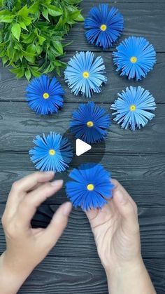 someone is holding blue paper flowers in front of a wooden table with green plants and greenery