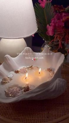 a white shell shaped candle holder on a table with pink flowers and a lamp in the background