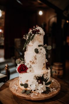 a white wedding cake with red flowers and greenery sits on a wooden platter