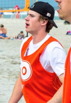 two men in orange vests standing on the beach with people in the water behind them