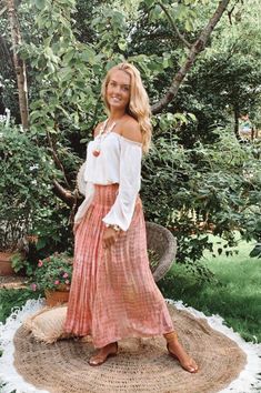 a woman standing on top of a rug in the middle of a garden wearing a white blouse and pink pleated skirt