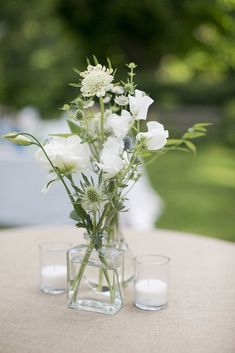 two vases filled with white flowers sitting on top of a table next to candles