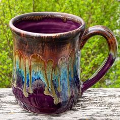 a purple and blue coffee mug sitting on top of a wooden table next to trees