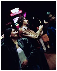 a man and woman standing next to each other on the street at night with neon signs in the background