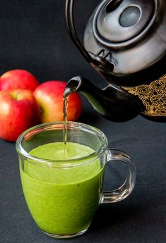 a glass cup filled with green liquid next to apples