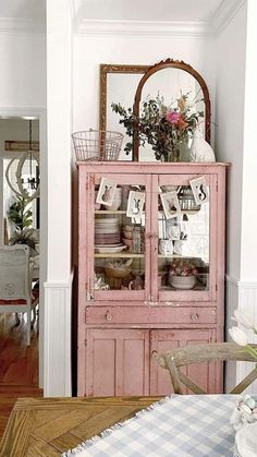 a pink china cabinet with flowers on top in a white walled dining room, next to a wooden table and chairs