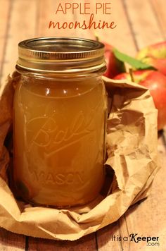 an apple pie in a mason jar on a wooden table with apples behind it and text overlay that reads how to make apple pie moonshine