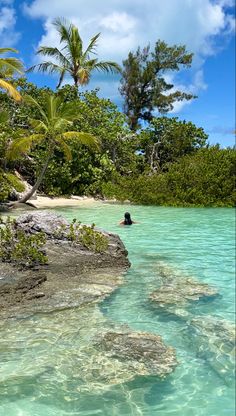 a person swimming in the ocean near some rocks and palm trees, with blue water