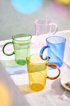 three different colored glass mugs sitting on top of a white cloth covered tablecloth