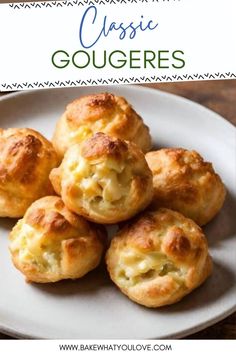 A plate of Gougeres served on a wooden table, with a piece of cheese visible to the side. Gourges Cheese Puffs, Appetizers With Gruyere Cheese, Gougeres Recipe Ina Garten, Gouda Cheese Ball Recipes, Gouda Cheese Recipes Appetizers, Swiss Cheese Appetizers, Gouda Appetizers, Recipes With Gouda Cheese