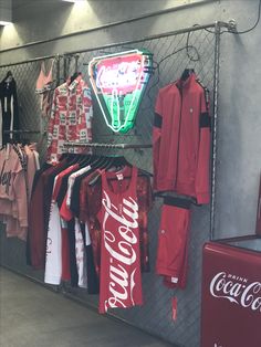 a coca - cola neon sign is hanging on the wall next to several shirts and coolers