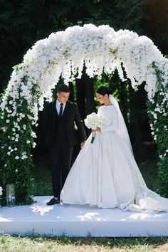 a bride and groom walking down the aisle under an arch with white flowers on it