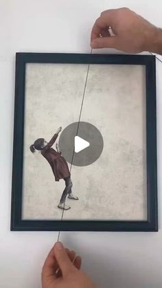 a person holding up a framed painting with a string attached to it and another hand reaching for the frame