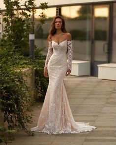 a woman in a long sleeved wedding dress standing on a sidewalk with greenery