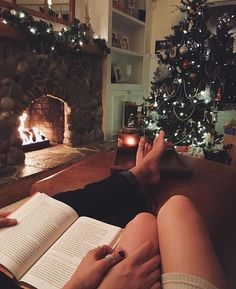 a person sitting on a couch reading a book in front of a christmas tree and fireplace