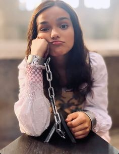 a young woman sitting at a table with her hand on her face and holding a chain around her neck