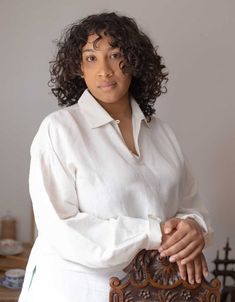 a woman with curly hair is posing for a photo in front of a wooden chair