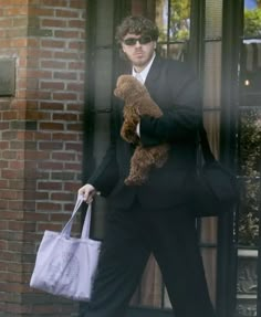 a man in a black suit carrying a brown teddy bear and a white tote bag