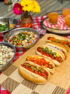 hot dogs on buns with toppings sit on a wooden cutting board next to bowls of salad