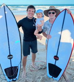 a man holding a baby and standing next to two surfboards on the beach,