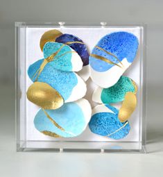 some blue and gold painted rocks in a clear box on a white table with silver foil