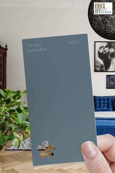 a person holding up a blue paint swatch in front of a living room with pictures on the wall