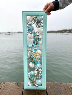 a person holding up a frame with seashells and starfish on it in front of the water