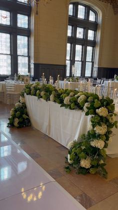 a table with white flowers and greenery on it in front of large windows at a wedding reception