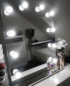 a mirror with lights on top of it in front of a bed and vase filled with flowers