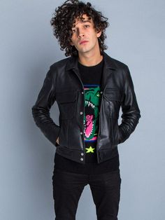 a man with curly hair wearing a black leather jacket and jeans standing in front of a gray wall