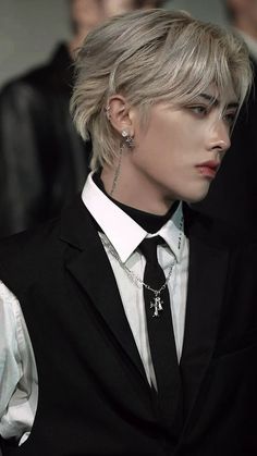 a man with blonde hair wearing a black suit and white shirt, silver necklace and earrings