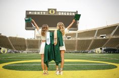 two women in green graduation gowns standing on a football field with their hands up