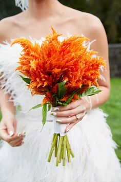 a woman in a white dress holding a bouquet of orange flowers and feathers on her wedding day