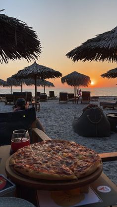 a pizza sitting on top of a wooden table next to the ocean with straw umbrellas