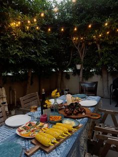 an outdoor dining table with food on it and lights strung over the tables in the background