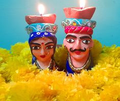 two colorfully painted masks with candles in their mouths, surrounded by bright yellow feathers