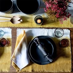 the table is set with black plates, silverware and vases on yellow placemats