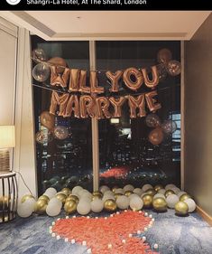 balloons and streamers spell out the word will you marry me in front of a window