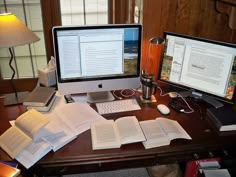 two computer monitors sitting on top of a desk next to an open book and lamp