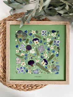 a green framed artwork with flowers and leaves on the wall next to a wicker basket