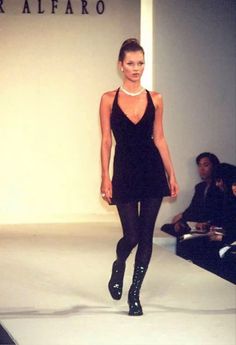 a model walks down the runway in a short black dress and high heeled boots