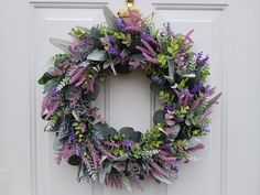 a wreath with purple flowers and greenery hangs on a white front door, next to a bell