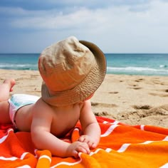 a baby laying on top of a beach next to the ocean with a hat on