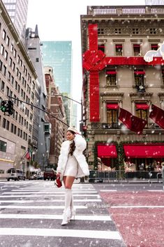 a woman in white is crossing the street with snow falling on her and buildings behind her