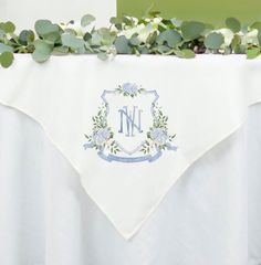 a white table cloth with an initial monogram on it and greenery around the edges