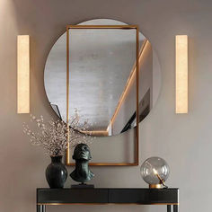 a mirror and vase with flowers on a table in front of a wall mounted light